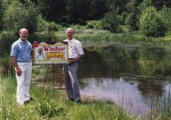 Two men standing together at the edge of the pond in summer, one holding a book.