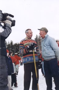 Two men standing on the ice mid-interview, one holding a microphone.